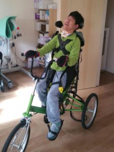 A boy in a green jumper on a Tomcat trike in a medical room.