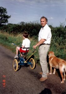 Bob and Tom on a road. Tom is looking away from the camera, facing forwards on the trike, and Bob has turned back to look at the camera. He is holding the Carer Control arm. There is a dog on his right, also facing up the road.