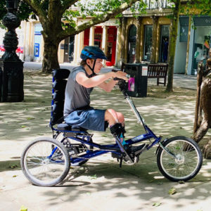 A man on a Tomcat trike in a town centre. He is perpendicular to the camera, looking at something off-screen.