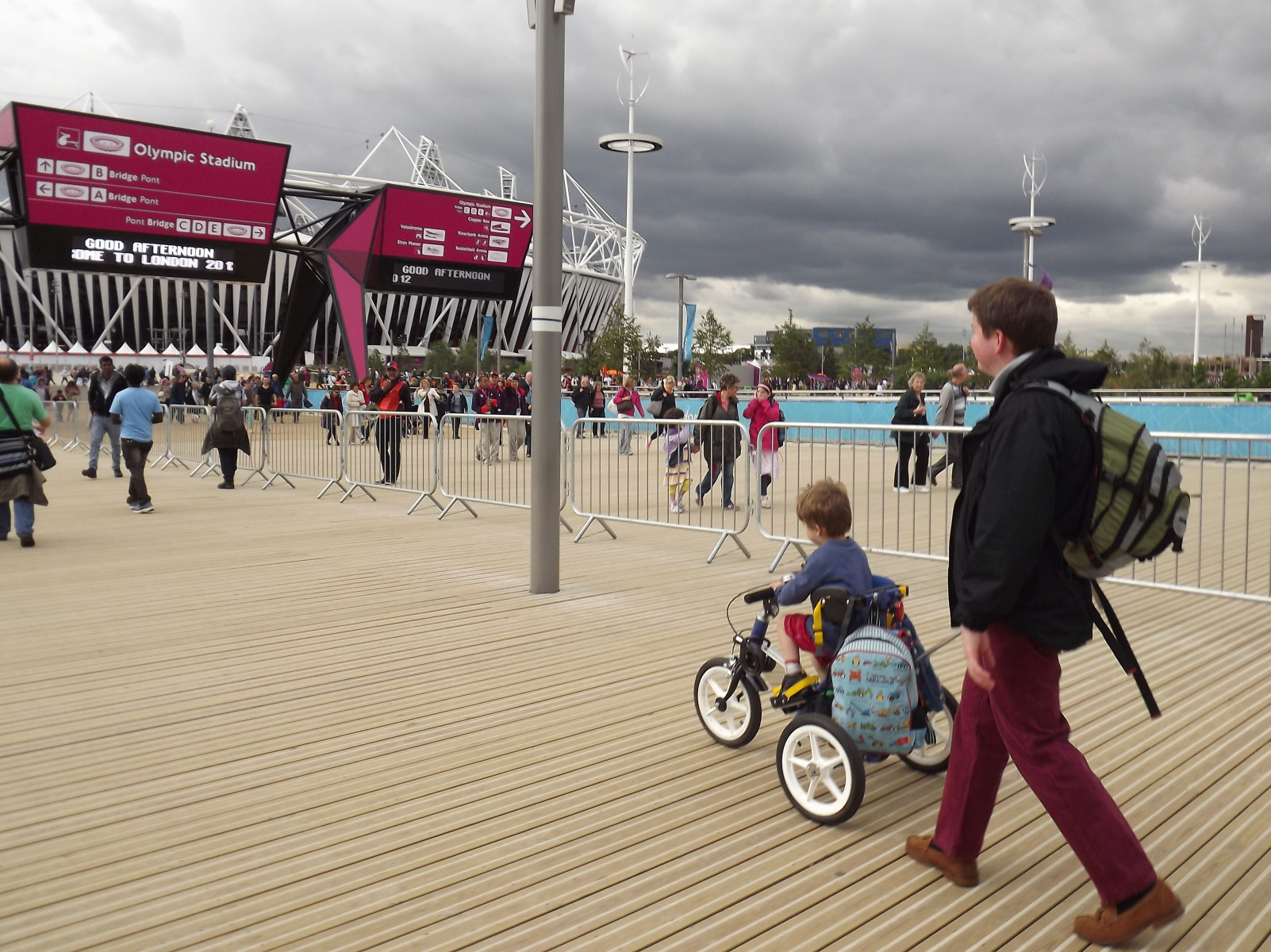 A child on a Tomcat Tiger cycling along a boardwalk up to the Olympic stadium. There is an adult behind holding the Carer Control.