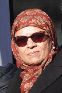 An older woman in a red patterned headscarf and sunglasses. She is wearing a black coat with lapels.