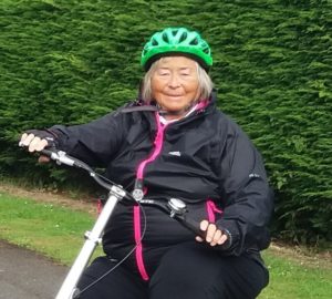 A woman in a black raincoat and green cycling helmet, sat on a trike in front of a large green hedge.