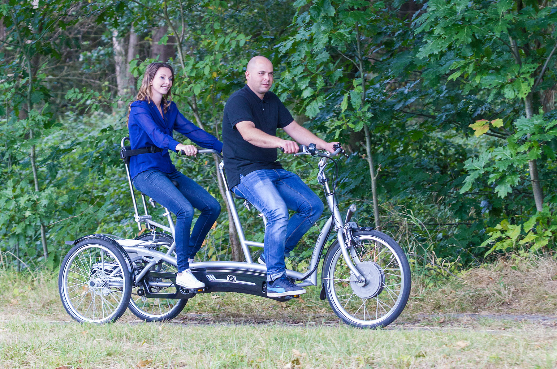 A man and woman on a Twinny Plus Tandem, cycling along a park path with trees behind them.