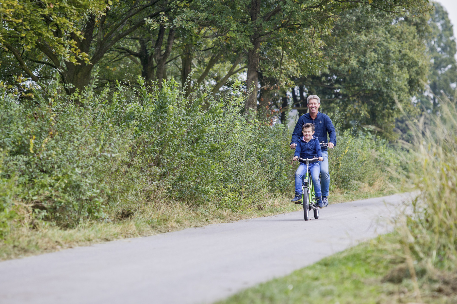 An adult and child riding a Kivo Tandem bike down a cycle path with hedges and trees on either side.