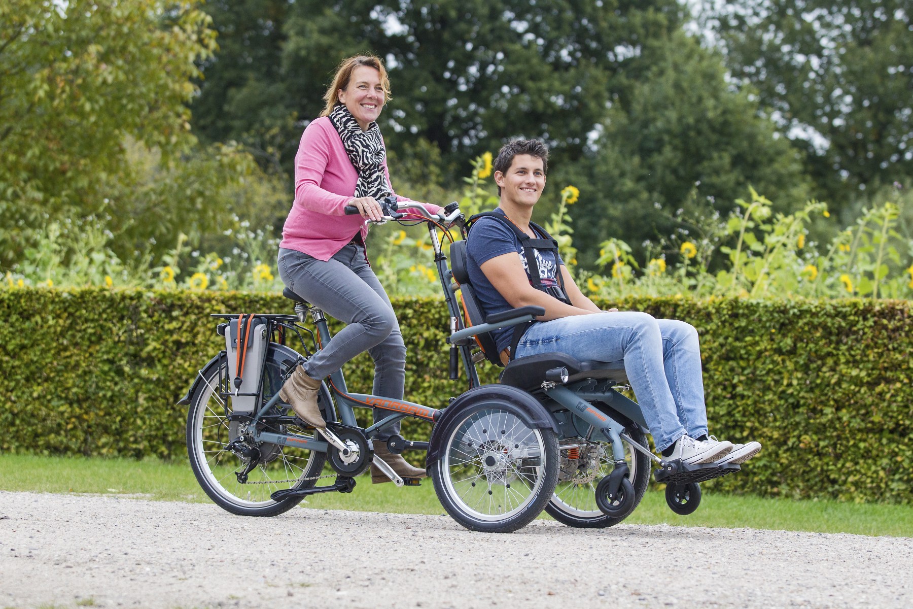 Two people on a Van Raam OPair bike, cycling through a park. They are smiling at something beyond the camera.