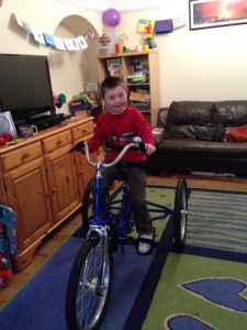 Joe, in his living room, on his Tomcat Trike. The trike is blue, and Joe is wearing a red shirt and dark grey trousers. He is smiling at the camera.