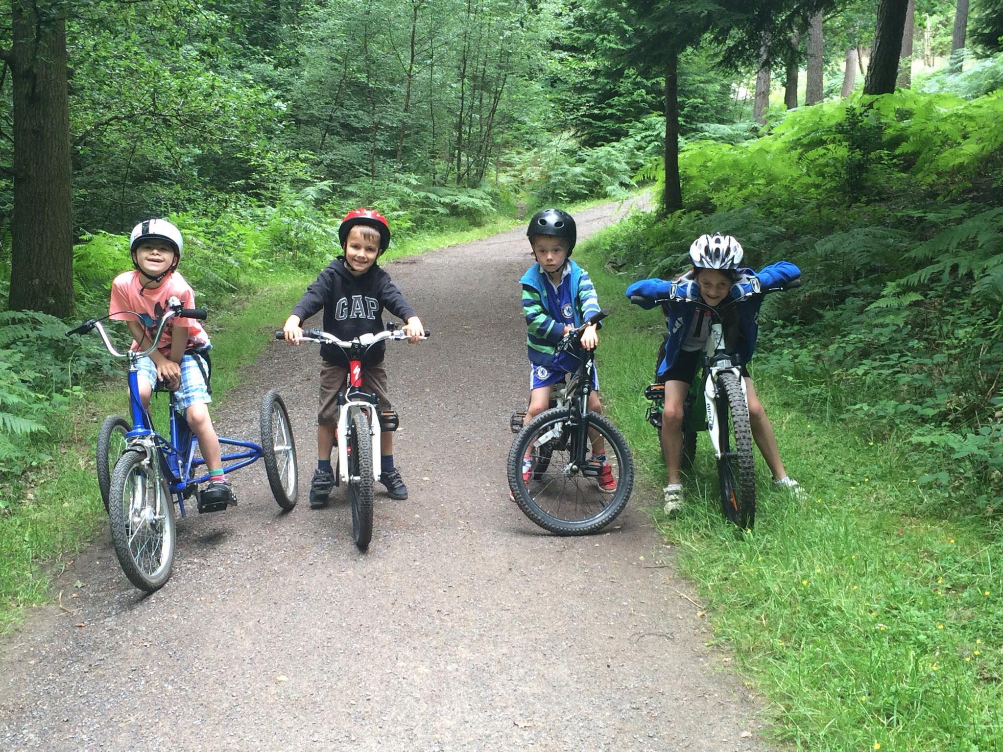 Four children on a forest cycling trail, all with bikes. Joe is on the left on a Tomcat trike. They are all smiling at the camera.