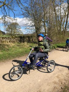 Nathan on his Bullet trike, on a cycling track in the countryside. He is wearing a green puffer jacket and a blue cycling helmet.