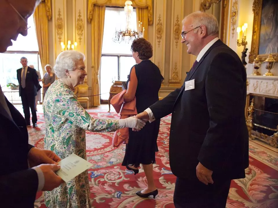 Bob, in a black suit with a nametag, shaking hands with the Queen.