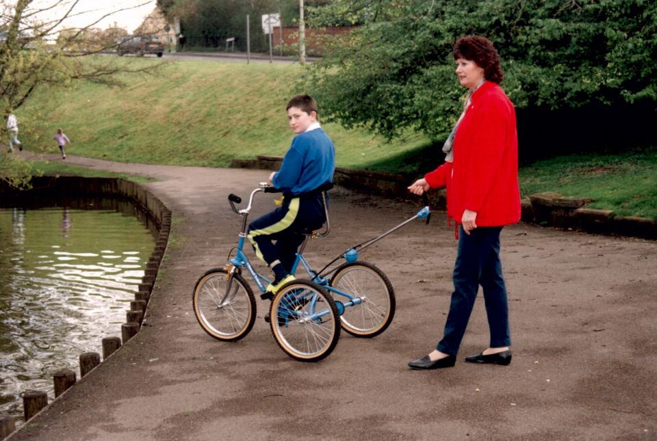 Tom, on a trike, by the lake with Anna, his mum. Tom is facing the lake, and Anna is holding the Carer Control handle to demonstrate its effectiveness.