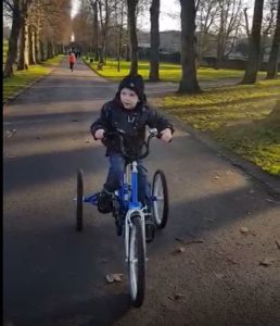 Charlie on his blue Tomcat trike, cycling down a tarmac path with rows of trees either side.