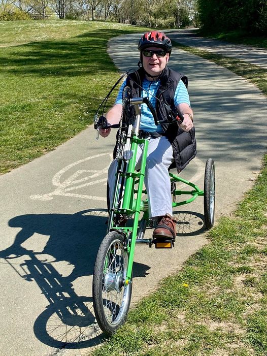 A person on a green Rotor Bullet Tomcat trike, on a cycle path in a park. They are wearing a helmet and smiling at the camera.