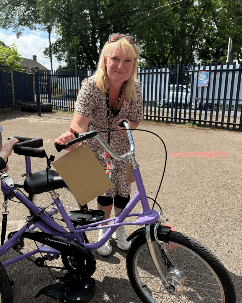 Photo of a smiling lady stood next to a purple Tomcat Fizz Tricycle that was donated to her school by Tomcat.