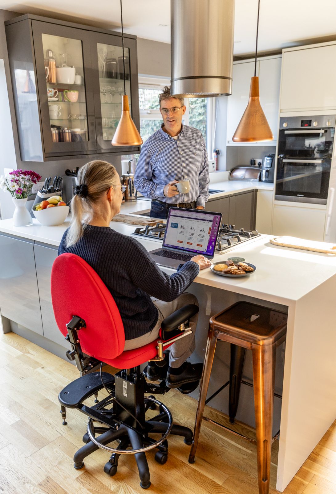 Woman using a Tomcat Solo Seat chair at a kitchen island, browsing the Tomcat website on a laptop while talking to a man