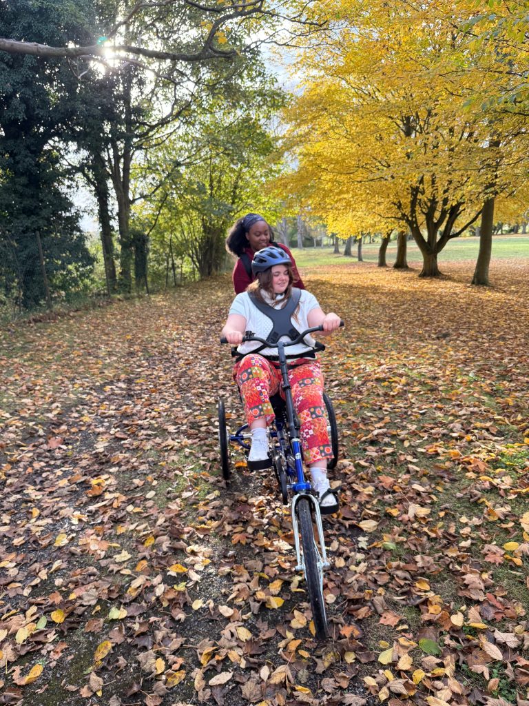 Sapphi on her Tomcat Apprentice Bullet Tricyle. Surrounded by yellow trees and fallen leaves. She is being pushed along by her carer using the Tomcat 'Carer Control Handle'.