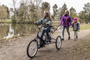 Child riding a purple Tomcat trike along a lakeside path with a woman and young child walking behind
