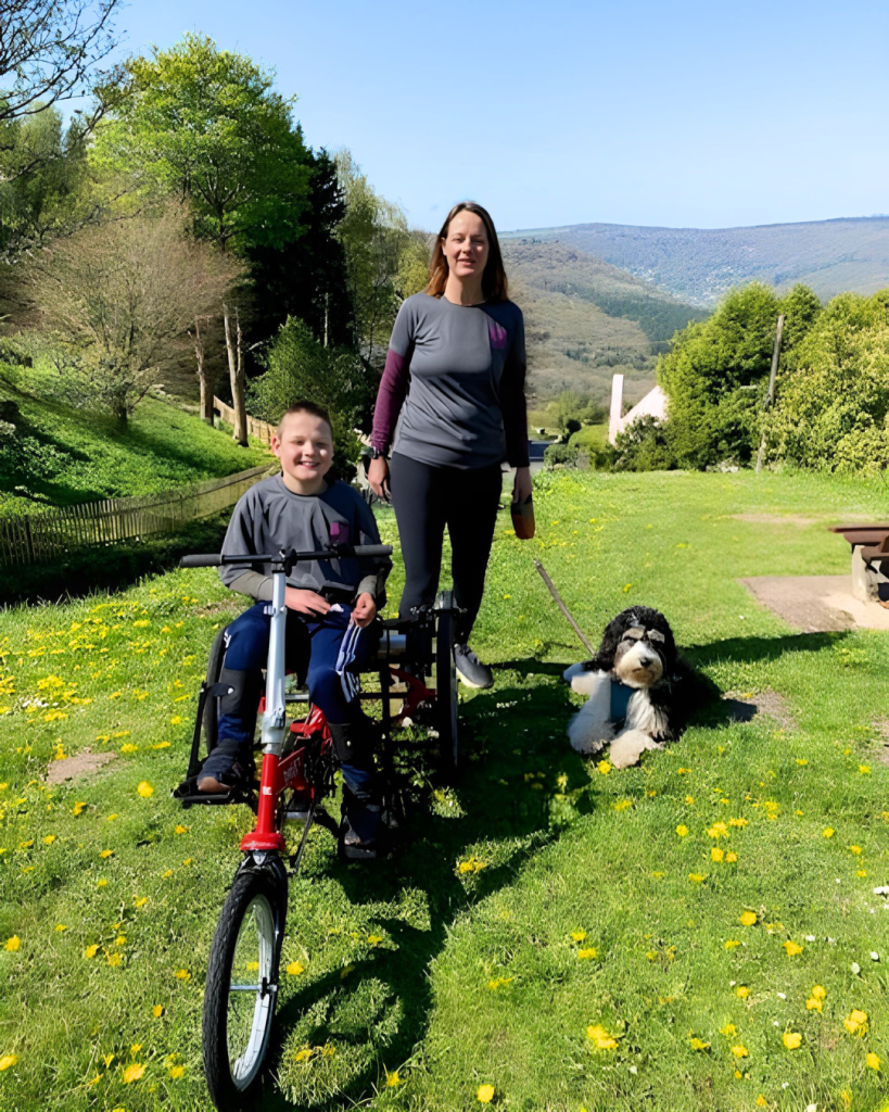 Boy riding a red Tomcat Apprentice Arrow adaptive trike with supportive seating while his mother walks beside him and their dog rests on the grass in a sunny countryside setting.