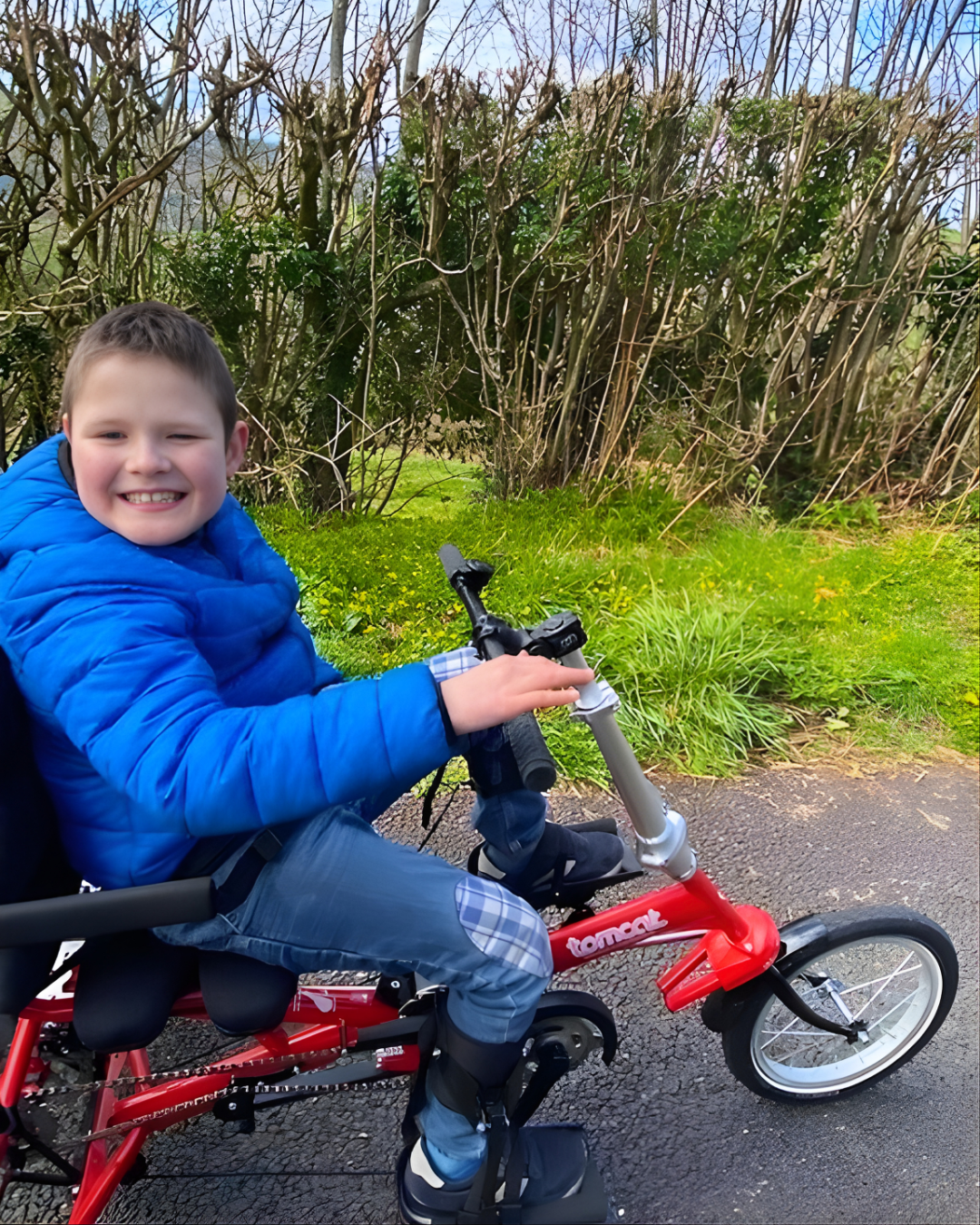 Oscar smiling while riding a red Tomcat Apprentice Arrow adaptive trike with supportive seating and leg supports along a countryside path.