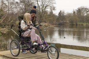 Teenage girl riding a purple Tomcat trike with Carer Control on a lakeside jetty, smiling with a woman guiding from behind